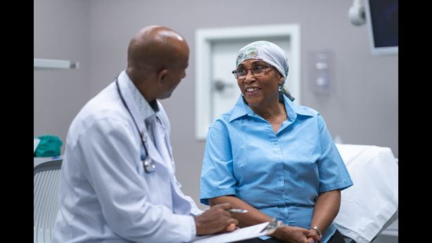 A black senior woman with cancer is at a medical consultation. She is wearing a scarf on her head to hide her hair loss. The doctor is a black man. The patient and doctor are sitting next to each other on an examination table in a medical clinic. The doctor is asking the patient questions and taking notes on a clipboard. The patient is smiling with hope and gratitude.